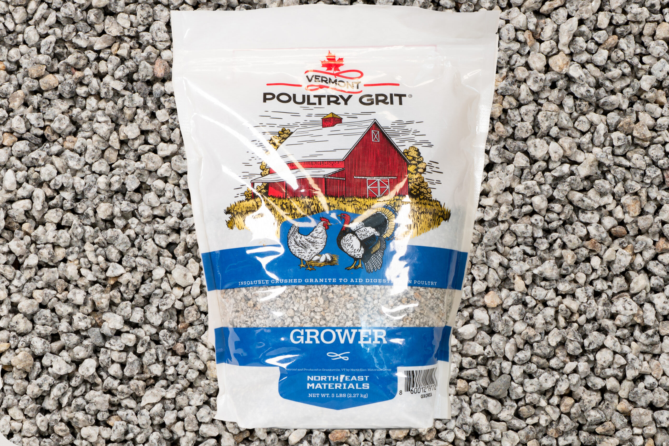 A bag of Vermont Poultry Grit Grower sits on gravel, featuring an illustration of a red barn with two chickens in front. This poultry grit, made from crushed granite, helps aid digestion in poultry.