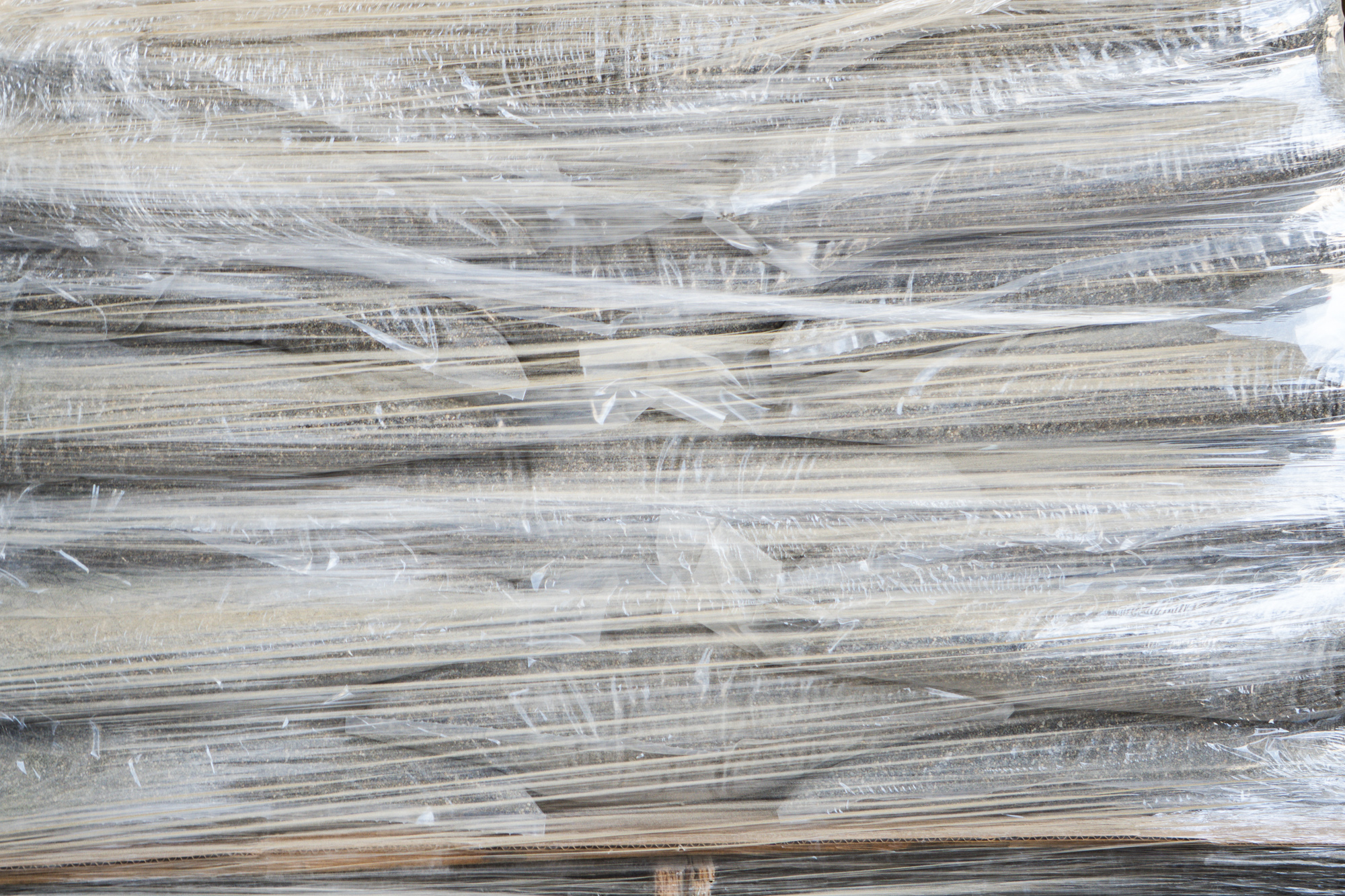 A close-up view of a stack of cardboard or wooden sheets wrapped tightly in clear plastic stretch wrap, creating a crinkled, shiny texture across the surface—ready for secure shipping.