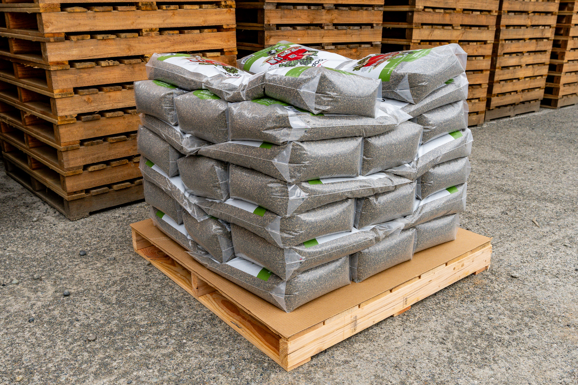 A stack of plastic bags filled with granular material is neatly arranged on a wooden pallet, ready for shipping, and surrounded by additional wooden pallets in the background on a concrete surface.