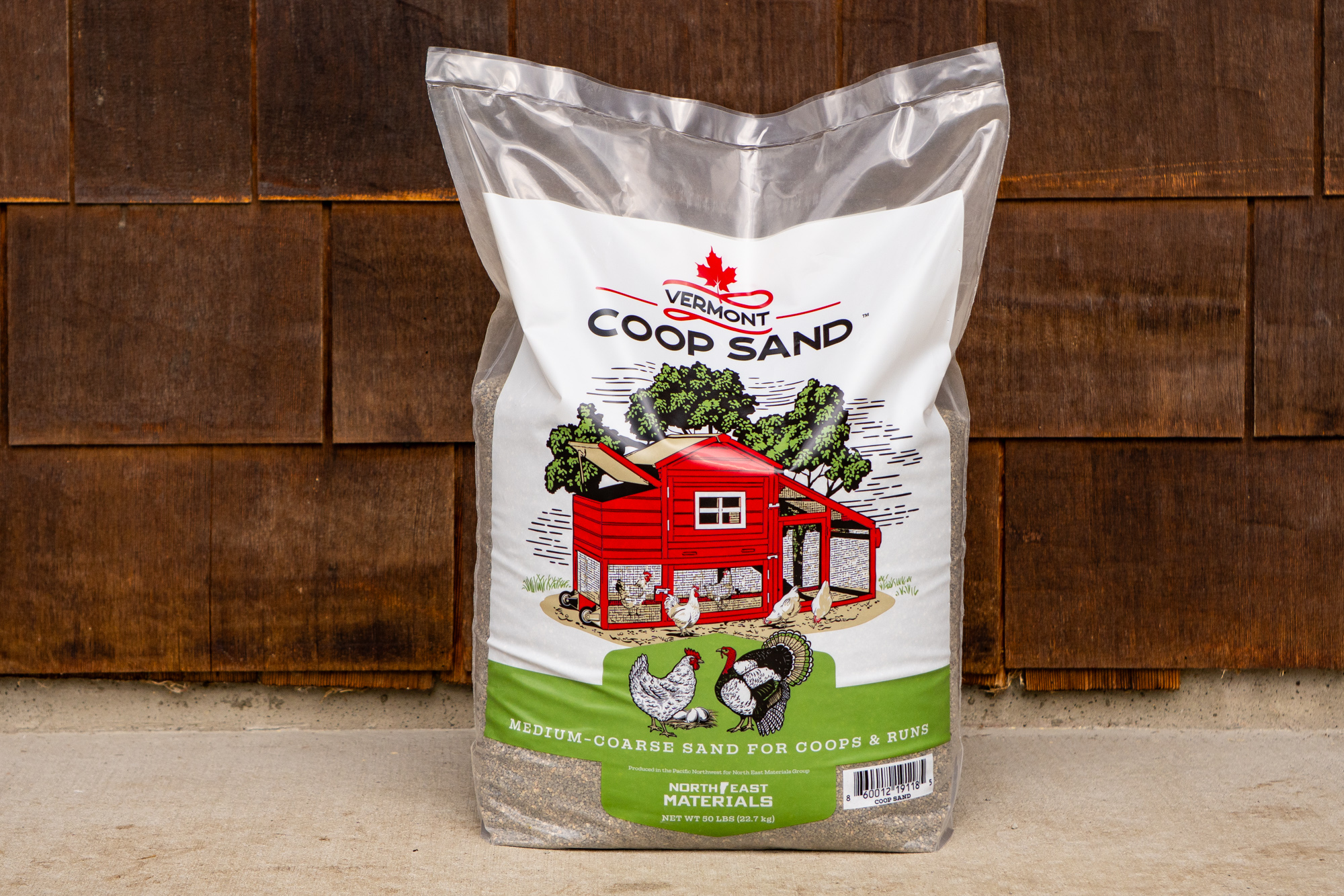 A clear plastic bag labeled Vermont Coop Sand, featuring a red chicken coop and three chickens, contains premium medium-coarse coop sand for coops and runs, resting on a concrete surface in front of a wooden wall.